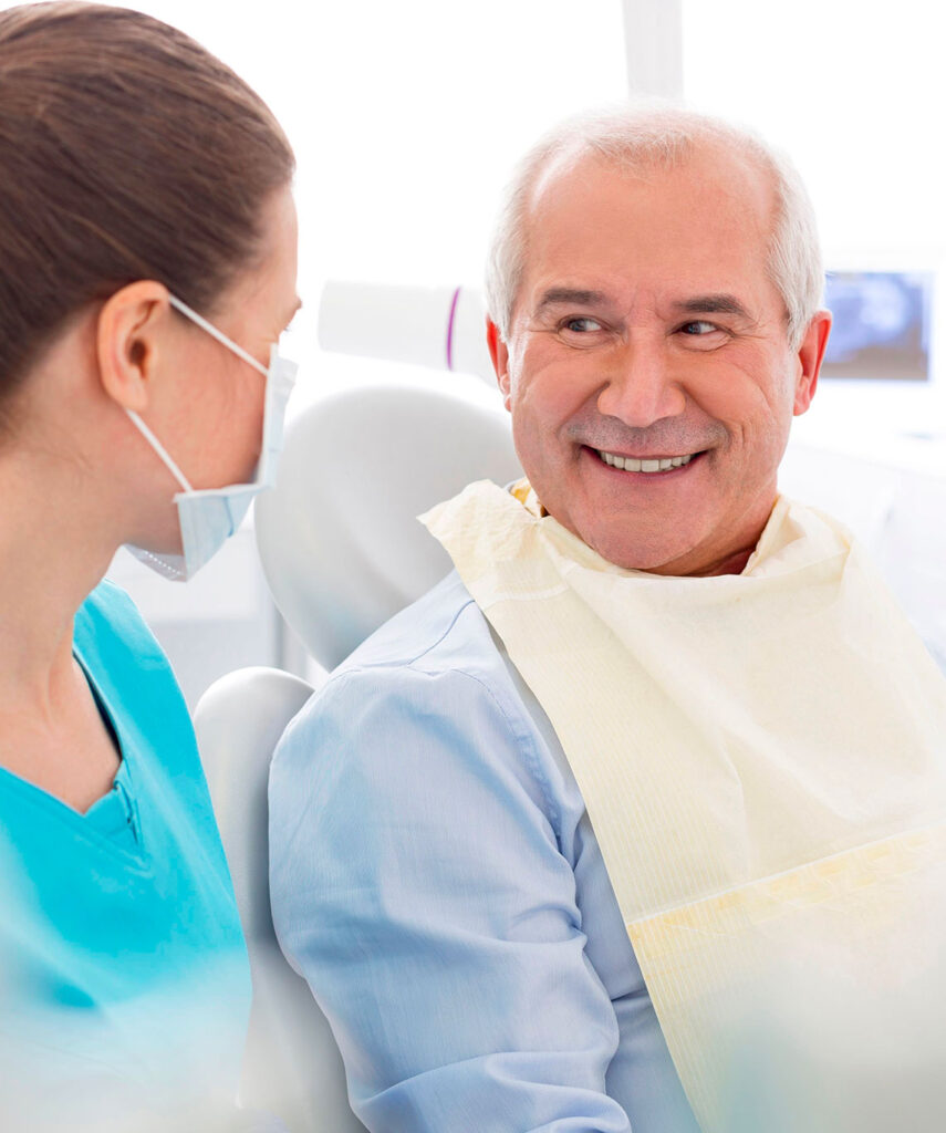 Dans un cabinet du Centre dentaire Deslauriers Pissardo Jacques, une assistante en blouse bleue s'adresse à un patient souriant assis dans un fauteuil dentaire. L'arrière-plan évoque un espace moderne et lumineux avec un écran visible.